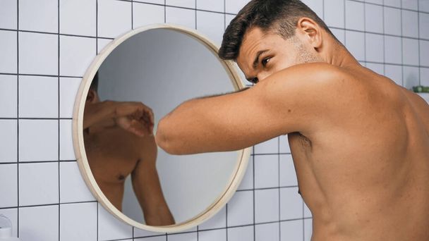 Мужчина без рубашки нюхает себя возле зеркала в ванной - Фото, изображение