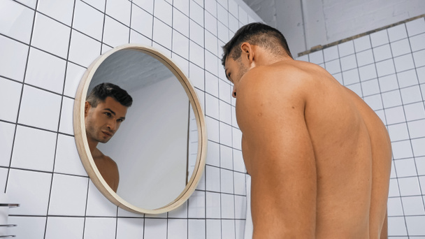 άποψη χαμηλή γωνία του νεαρού άνδρα shirtless κοιτάζοντας καθρέφτη στο μπάνιο - Φωτογραφία, εικόνα