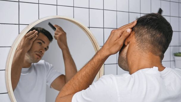 νεαρός άνδρας βούρτσισμα των μαλλιών με χτένα κοντά στον καθρέφτη στο μπάνιο, θολή φόντο - Φωτογραφία, εικόνα