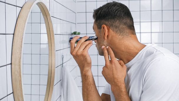 молодой человек бритья лицо с электрической бритвой возле зеркала в ванной комнате - Фото, изображение