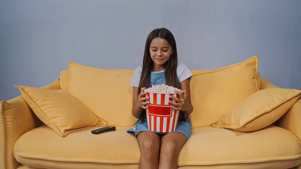 boldog gyerek nézi vödör popcorn, miközben ül a kanapén  - Fotó, kép