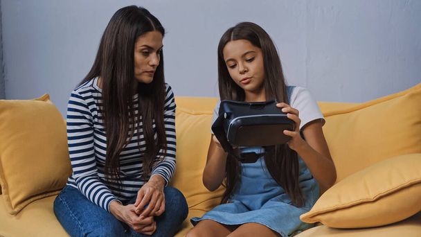 kid holding vr headset near mother sitting on couch - Photo, Image