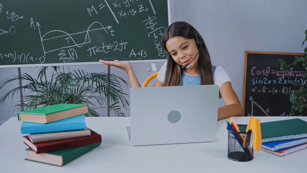gelukkig schoolkind in headset kijken naar laptop terwijl wijzend met de hand op schoolbord  - Foto, afbeelding