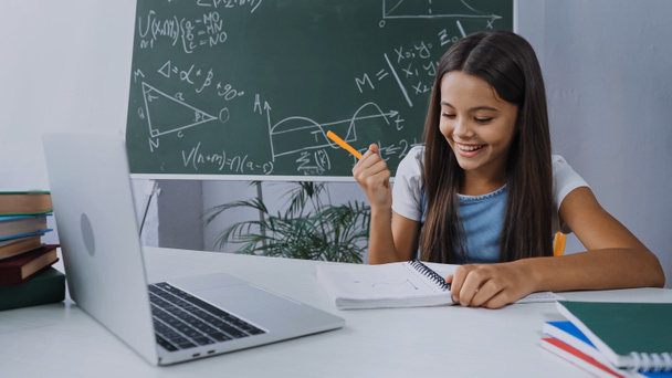 boldog lány kezében toll és nézi jegyzettömb mellett laptop az asztalon - Fotó, kép