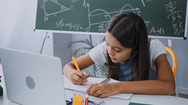 schoolkid in headset writing in notebook near laptop on desk - 写真・画像
