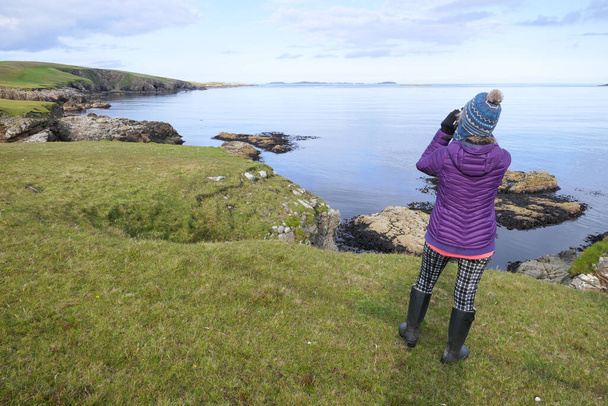 Una dama en ropa de invierno y sombrero bobble mira con prismáticos en un paisaje costero de rocas y mar.Shetland.UK - Foto, imagen