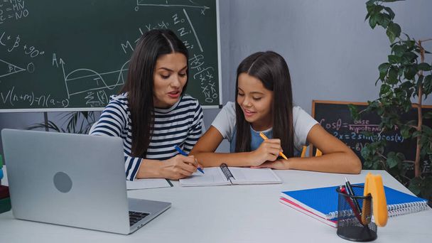мать и счастливая дочь делают домашнее задание возле ноутбука на столе - Фото, изображение