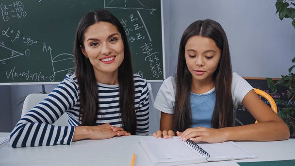 mother smiling near daughter doing homework at home - Photo, Image