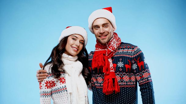 Smiling couple in sweaters and santa hats hugging on blue background - Фото, изображение