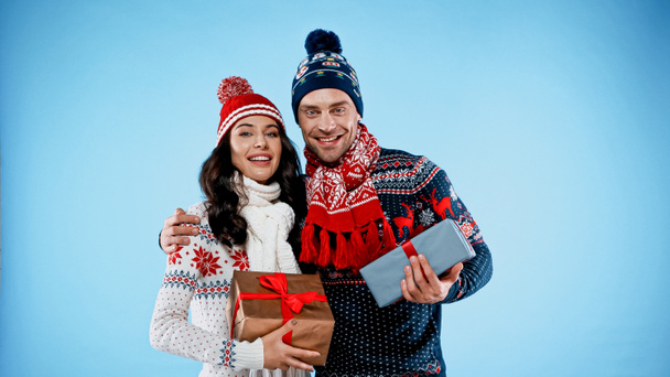 Χαμογελαστό ζευγάρι με πουλόβερ και καπέλα που κρατούν δώρα σε μπλε φόντο - Φωτογραφία, εικόνα