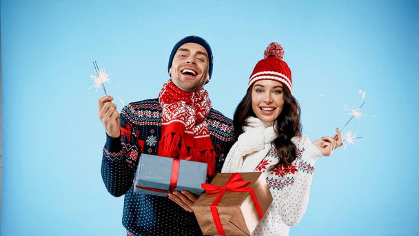 Fröhliches Paar in Pullovern mit Wunderkerzen und Geschenken auf blauem Hintergrund - Foto, Bild