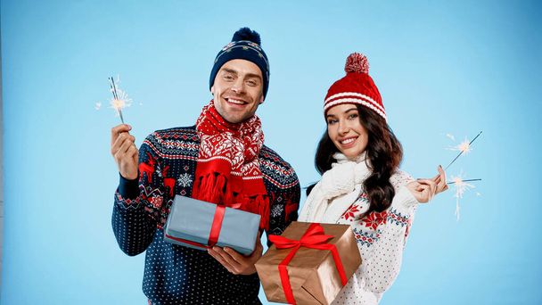 Couple in warm sweaters holding sparklers and gifts on blue background - Zdjęcie, obraz