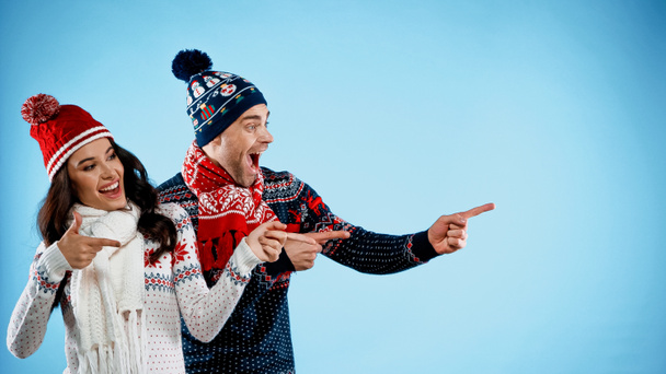 Cheerful couple in hats and sweaters pointing with fingers on blue background - Photo, Image