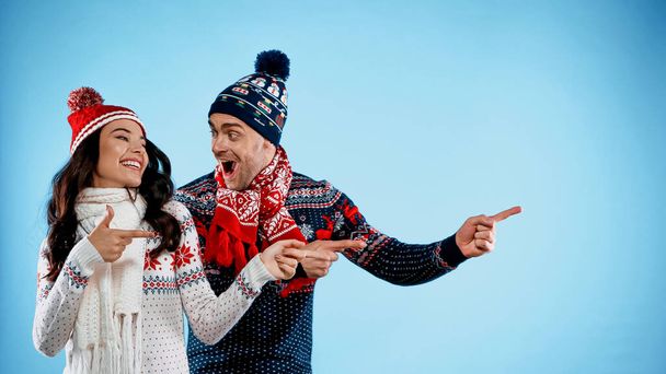 Excited man in sweater pointing with finger near smiling girlfriend on blue background - 写真・画像