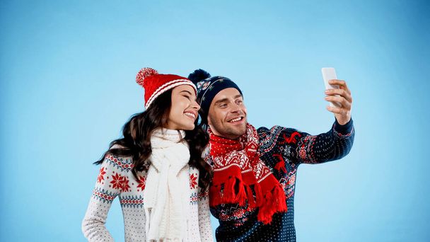 Szczęśliwa para w kapeluszach, swetrach i szalikach robi selfie na niebieskim tle - Zdjęcie, obraz