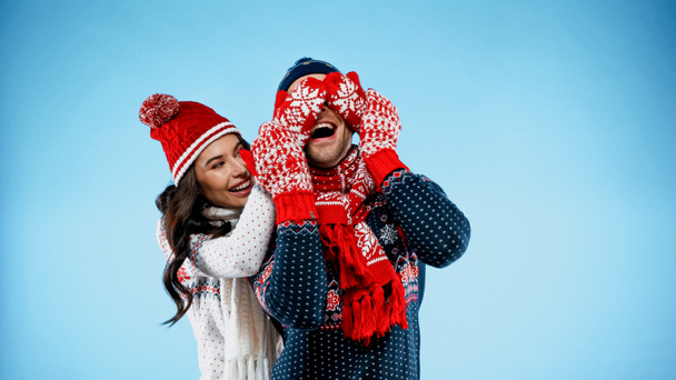 Cheerful woman in sweater and mittens covering eyes of boyfriend on blue background - Photo, Image