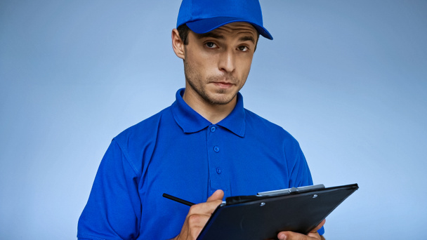 serious delivery man writing order on clipboard isolated on blue - Фото, изображение