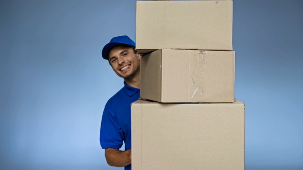 veselá dodávka muž drží kartonové krabice, zatímco se usmívá na kameru izolované na modré - Fotografie, Obrázek