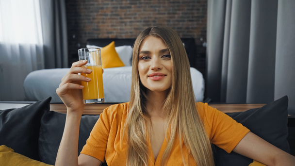 ευτυχισμένη νεαρή γυναίκα κρατώντας ένα ποτήρι χυμό πορτοκάλι στο δωμάτιο του ξενοδοχείου  - Φωτογραφία, εικόνα