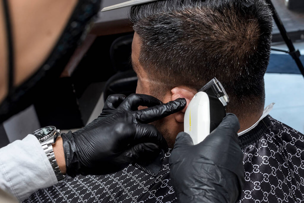 Holič s černými rukavicemi používá střihač na vlasy k ostříhání a ostříhání zákazníka. Klasický a módní účes. V holičství.. - Fotografie, Obrázek