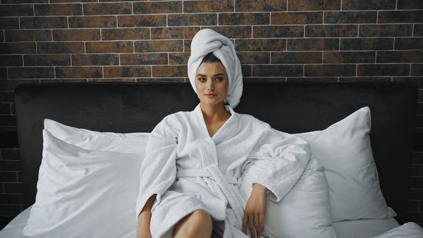 žena v bílém ručníku při pohledu na kameru, zatímco odpočívá na posteli v hotelovém pokoji - Fotografie, Obrázek