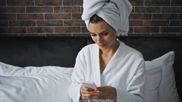 femme en serviette blanche regardant smartphone tandis que la messagerie dans la chambre d'hôtel - Photo, image