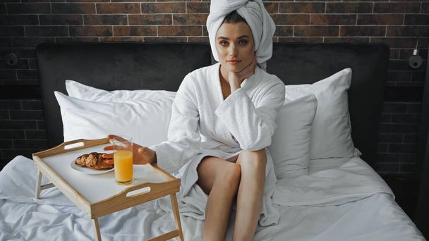 Frau in weißem Handtuch und Bademantel blickt in die Kamera neben Tablett mit Frühstück auf dem Bett - Foto, Bild