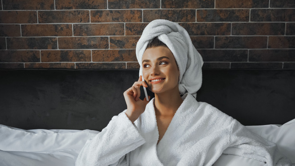Beyaz havluyla bornozlu mutlu kadın otel odasında akıllı telefondan konuşuyor. - Fotoğraf, Görsel