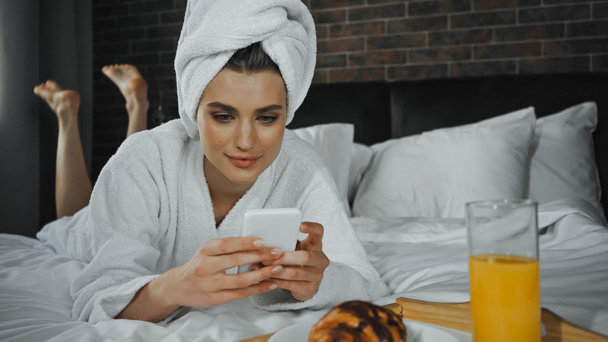 junge Frau in Handtuch mit Smartphone in der Nähe des Frühstücks auf Tablett im Hotelzimmer - Foto, Bild