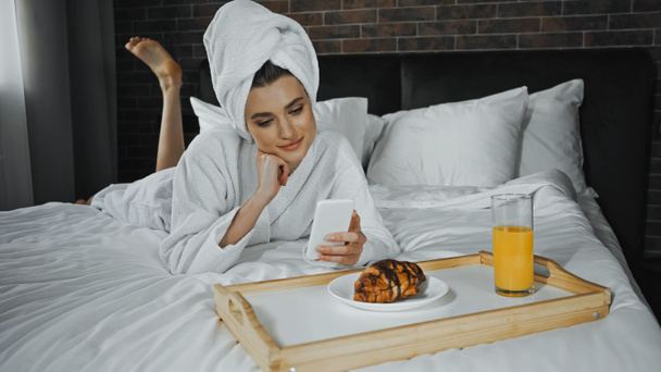 タオルとバスローブを着た若い女性ホテルの部屋のトレイで朝食の近くにスマートフォンを使って - 写真・画像