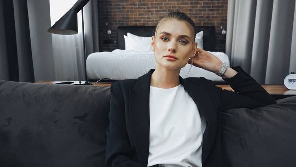 businesswoman in formal wear sitting on sofa and looking at camera in hotel room - Photo, Image