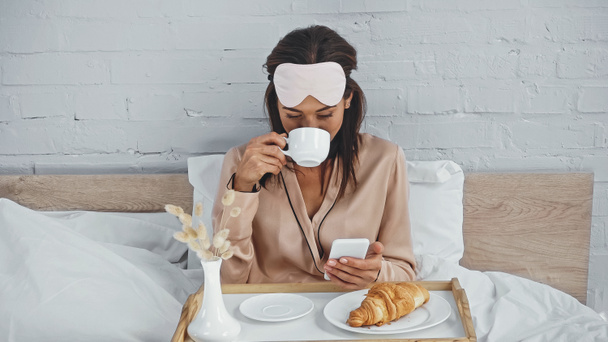 ブルネットの女性がコーヒーを飲み、ベッドの上でスマートフォンでメッセージ - 写真・画像