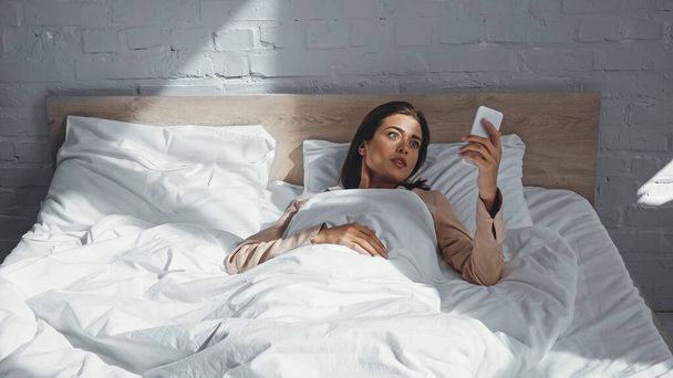 zaskoczona kobieta patrząc na smartfona leżąc w łóżku - Zdjęcie, obraz