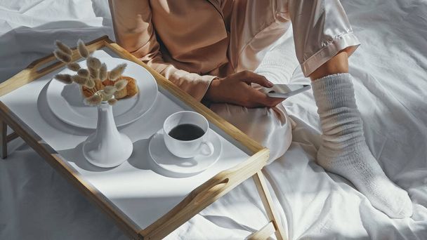 vista recortada de la mujer usando teléfono inteligente cerca de la bandeja con desayuno sabroso  - Foto, imagen