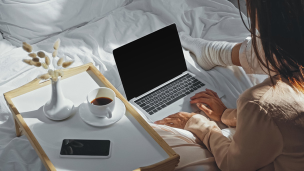 visão de alto ângulo da mulher segurando xícara de café perto do laptop com tela em branco na cama - Foto, Imagem