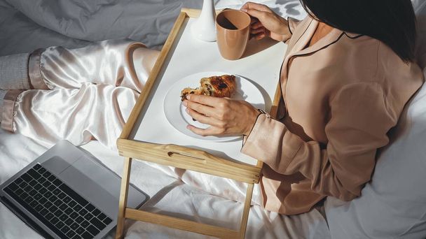 cropped view of woman holding cup of tea and croissant near laptop on bed - Photo, Image