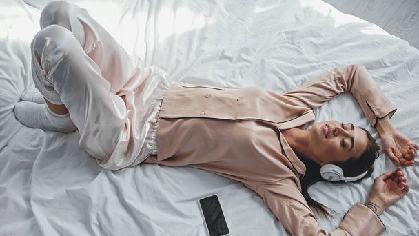vista ad alto angolo di donna felice in cuffie wireless sdraiato sul letto vicino smartphone con schermo bianco - Foto, immagini