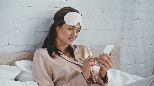 glückliche Frau mit Augenmaske textet auf Smartphone im Schlafzimmer  - Foto, Bild