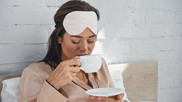 Kahve içerken elinde fincan ve fincan tabağı tutan göz maskeli genç bir kadın. - Fotoğraf, Görsel