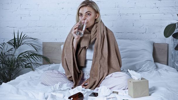 diseased woman drinking water while sitting in bed near medications - Foto, Imagem