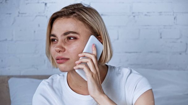 mujer joven enferma mirando hacia otro lado mientras habla en el teléfono móvil en el dormitorio - Foto, imagen