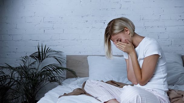 mujer enferma que oscurece la cara con las manos mientras estornuda en el dormitorio - Foto, imagen
