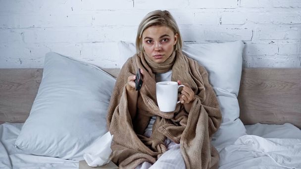 Kranke Frau sitzt mit Tasse Tee unter Decke vor dem Fernseher - Foto, Bild