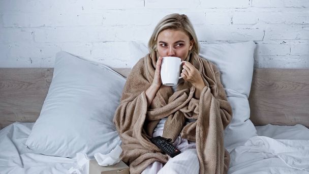 zieke vrouw drinken warme drank tijdens het kijken naar tv in de slaapkamer - Foto, afbeelding