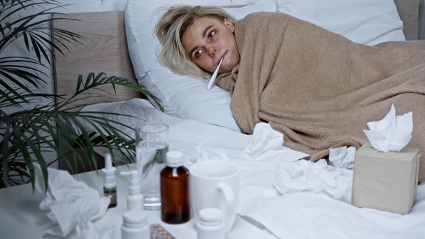 mujer enferma que mide la temperatura mientras está acostada en la cama cerca de medicamentos en primer plano borroso - Foto, Imagen