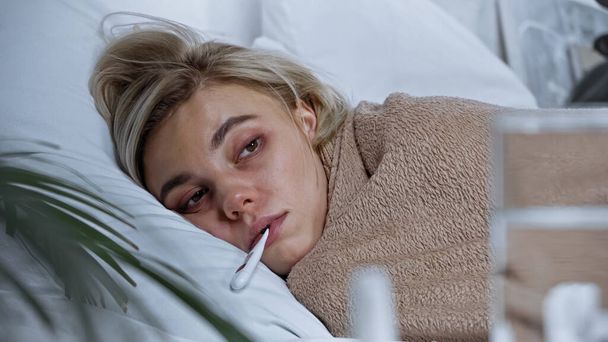 Kranke junge Frau misst Temperatur im Bett mit verschwommenem Vordergrund - Foto, Bild