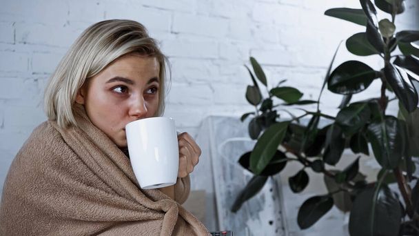 diseased blonde woman drinking warm tea while watching tv in bedroom - Photo, Image