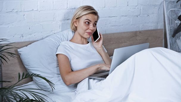 smiling freelancer talking on smartphone while using laptop in bedroom - Foto, afbeelding
