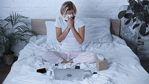 diseased freelancer sneezing into paper napkin near laptop and medications in bedroom - Φωτογραφία, εικόνα