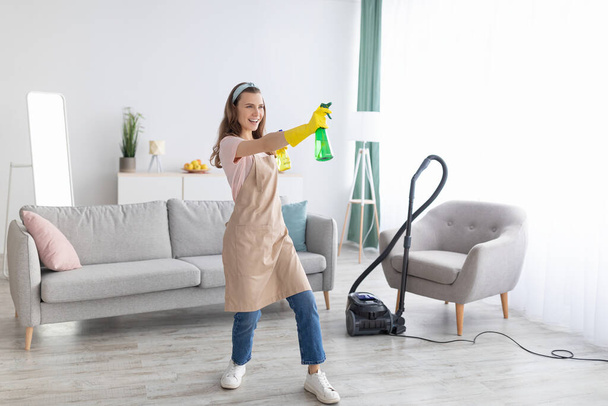 Gelukkig professioneel dienstmeisje met wasmiddel spray flessen in het midden van de schone woonkamer, kopieerruimte - Foto, afbeelding